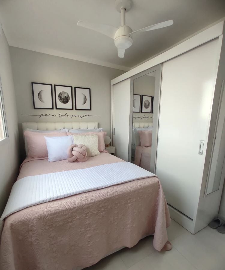 A imagem mostra uma decoração fácil para quarto com um ambiente claro e acolhedor. A cama tem uma colcha rosa claro e almofadas, contrastando com as paredes brancas. Acima da cama, há três quadros de arte com lua. Um armário com portas espelhadas amplia o espaço, sugerindo conforto e praticidade.