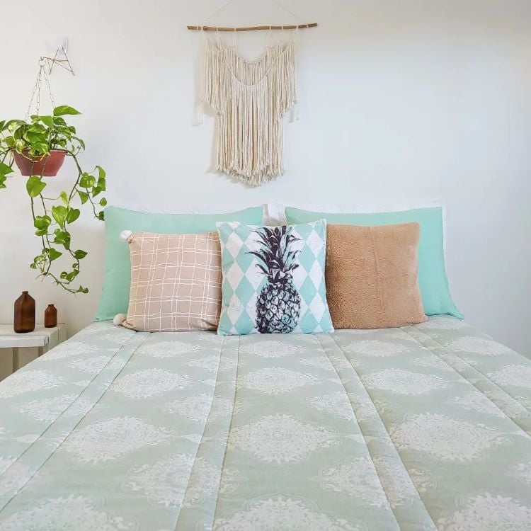 A imagem mostra uma decoração fácil para quarto com uma cama de frente para a câmera. A cama tem uma colcha geométrica verde e branca, três almofadas (uma com estampa de abacaxi), um enfeite de parede em macramê e um vaso suspenso com planta verde.