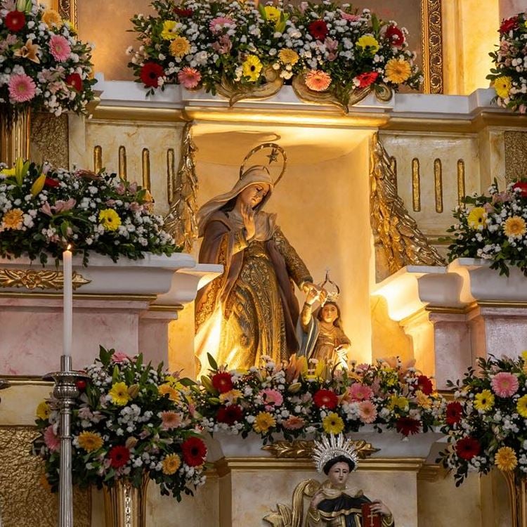 A imagem mostra uma estátua de uma figura feminina com um manto, provavelmente a Virgem Maria, segurando uma criança, possivelmente o menino Jesus. Eles estão cercados por flores coloridas e situados em um altar decorado com elementos dourados e velas acesas, criando um ambiente sagrado e reverente.