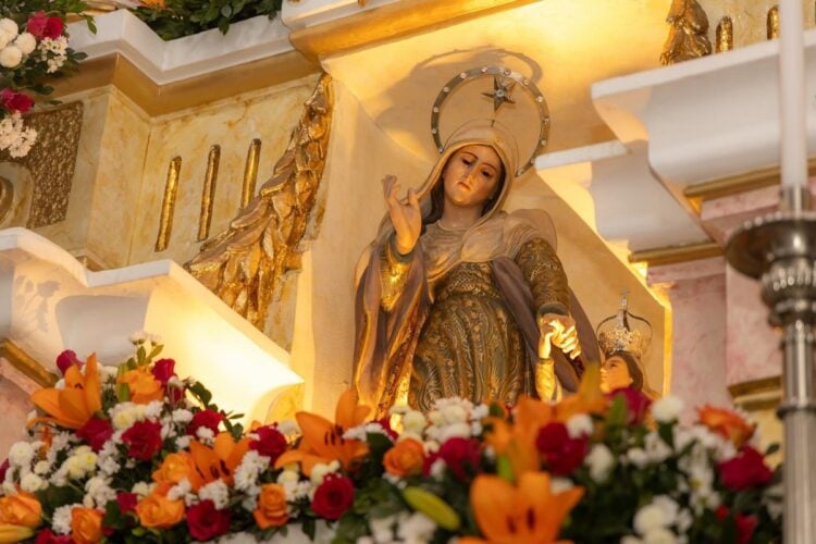 A imagem mostra uma estátua dourada de Sant’Ana, cercada por flores laranjas e amarelas. A figura possui asas e um halo, sugerindo uma representação celestial. Está posicionada em um altar com detalhes arquitetônicos que remetem a uma igreja, celebrando o Dia de Sant’Ana.