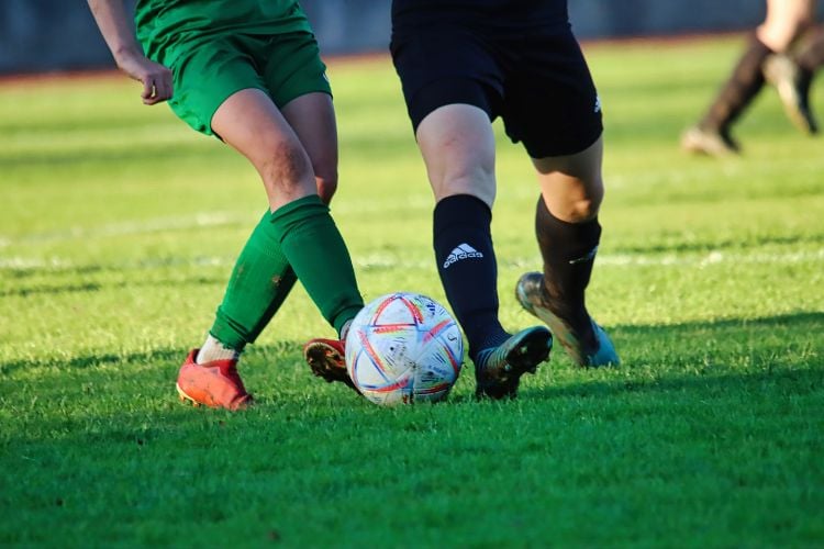 Pernas de duas pessoas disputando bola de futebol, uma de bermuda e meia verdes e outra de bermuda e meia pretas