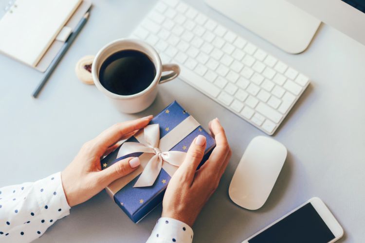 Pessoa de pele clara segurando embrulho azul de presente com laço dourado, em cima de mesa cinza com caneca de café, teclado branco e mouse branco