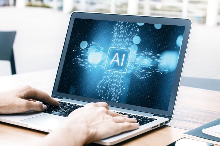 A imagem mostra um laptop sobre uma mesa com mãos digitando nele. A tela exibe uma representação gráfica de um conceito de inteligência artificial, com um cubo central rotulado “AI” e cercado por padrões de circuito e efeitos de luz, sugerindo conectividade digital e processamento de dados.