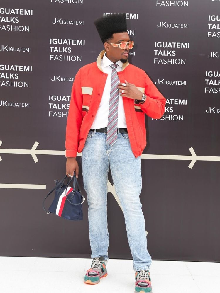 Matheus Pasquarelli usando jaqueta laranja, camisa branca, gravata colorida e calça jeans com parede com informações do Iguatemi Tallks Fashion atrás