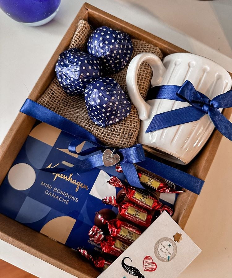 Uma caixa de presente com bombons, uma caneca e chocolates decorativos, todos com laços azuis, sugerindo um presente de Dia dos Pais.