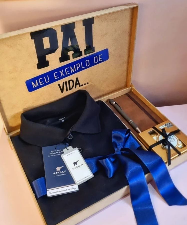 Caixa aberta contendo uma camisa polo azul dobrada, gravata azul, cinto e um cartão. Texto na tampa diz “PAI MEU EXEMPLO DE VIDA…”.