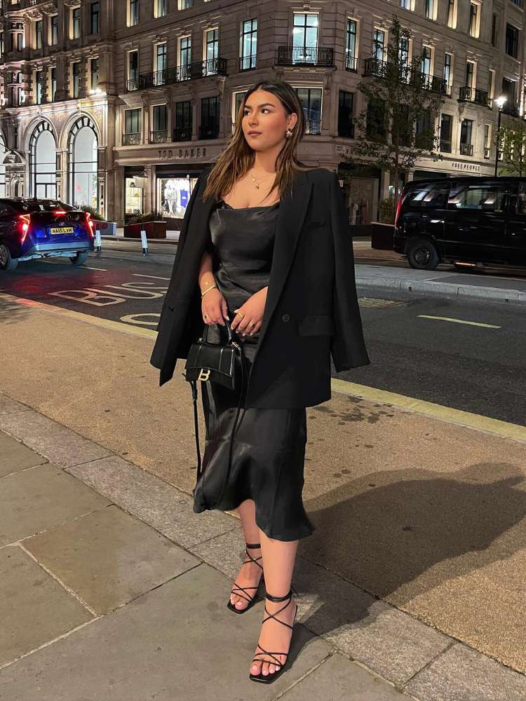 Mulher de pele clara usando vestido midi preto de cetim com blazer preto nos ombros, bolsa preta e sandália