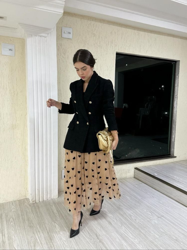 Mulher de pele clara com blazer preto fechado sobre vestido midi bege com corações pretos e bolsa dourada