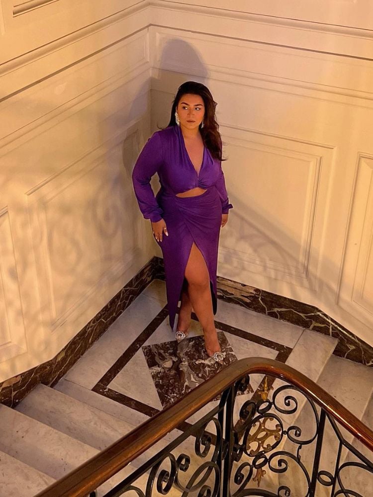 Mulher de pele morena em escada usando vestido midi roxo, com fenda lateral, decote e recorte na barriga 