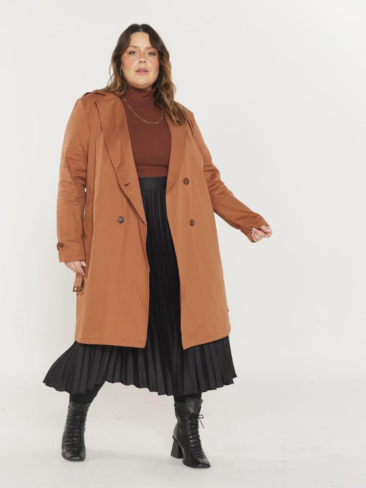 Mulher de pele clara usando blusa marrom, saia midi preta dratpeada, bota preta e trench coat marrom claro