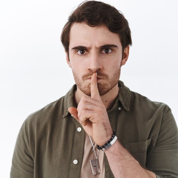 Homem de pele clara usando blusa marrom com semblante bravo e dedo indicador sobre a boca, em sinal de silêncio