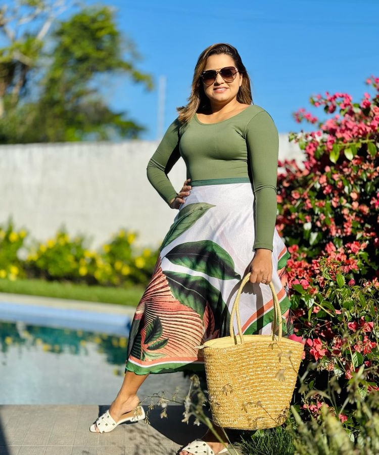 A imagem retrata uma pessoa usando um conjunto de moda evangélica: uma blusa de manga longa verde e uma saia midi estampada com folhagens em tons de verde, branco e rosa. Ela segura uma bolsa de palha e está ao lado de flores coloridas, sob a luz solar.