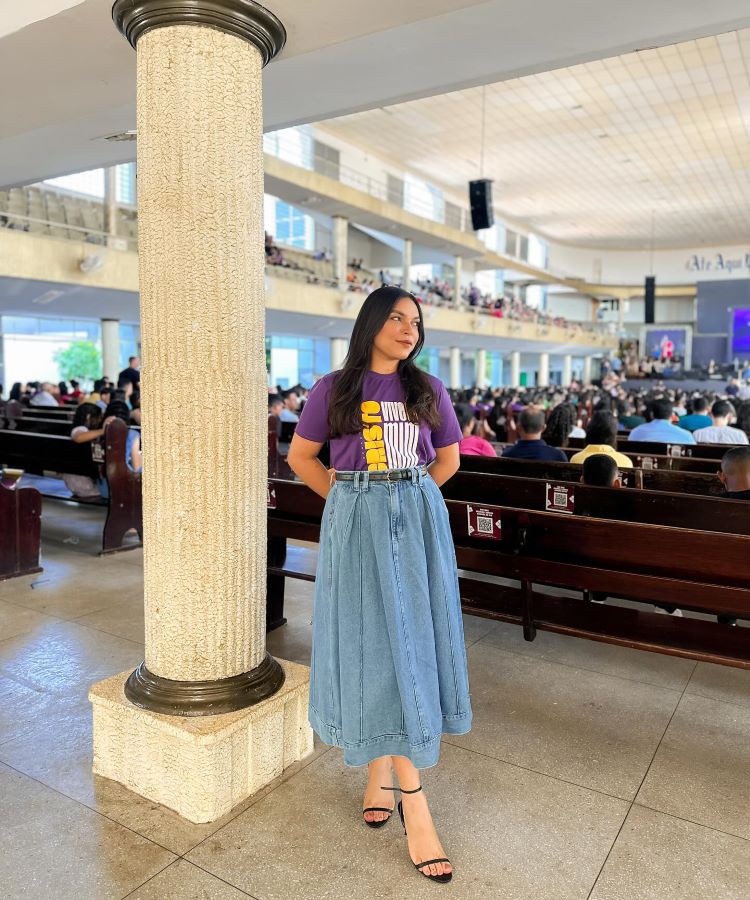 A imagem mostra uma mulher em uma igreja lotada, vestindo uma camiseta roxa com estampa "Cristo" e uma saia longa jeans, estilo moda evangélica. Ela está de pé, ao lado de uma coluna, com sandálias pretas de salto. Ao fundo, há muitas pessoas sentadas nos bancos.