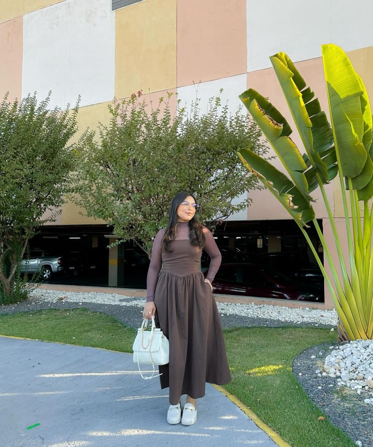 A pessoa na foto veste um elegante vestido marrom comprido, com mangas longas e gola alta. Ela segura uma bolsa branca e usa sapatos brancos. Ao fundo, há vegetação e parte de um estacionamento sob um edifício. A aparência é adequada para o estilo da moda evangélica.