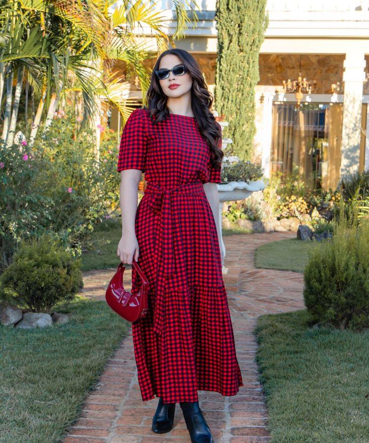 A pessoa na imagem veste um elegante vestido xadrez vermelho e preto, com mangas curtas bufantes e comprimento midi. Ela segura uma bolsa vermelha e usa botas pretas, enquanto o cenário ao fundo inclui um gramado bem cuidado e a entrada de uma casa. A moda evangélica valoriza peças modestas e sofisticadas.