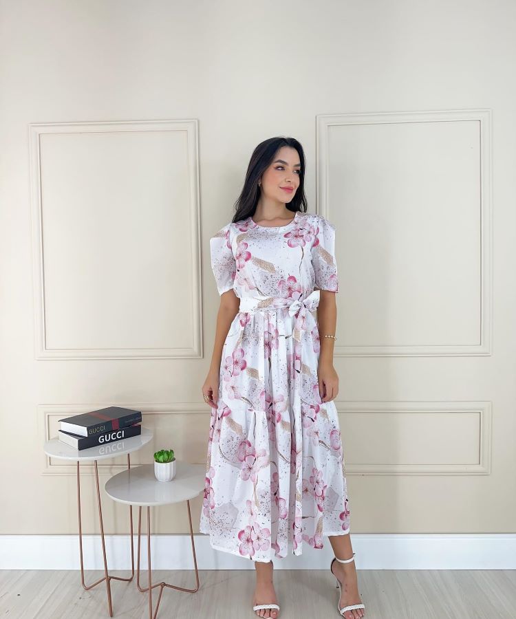 A pessoa na foto usa um vestido branco com estampa floral rosa, representando a moda evangélica. O vestido tem mangas curtas e cintura marcada. Ela está em pé em frente a uma parede clara com painéis decorativos, ao lado de uma mesa com livros e uma planta.