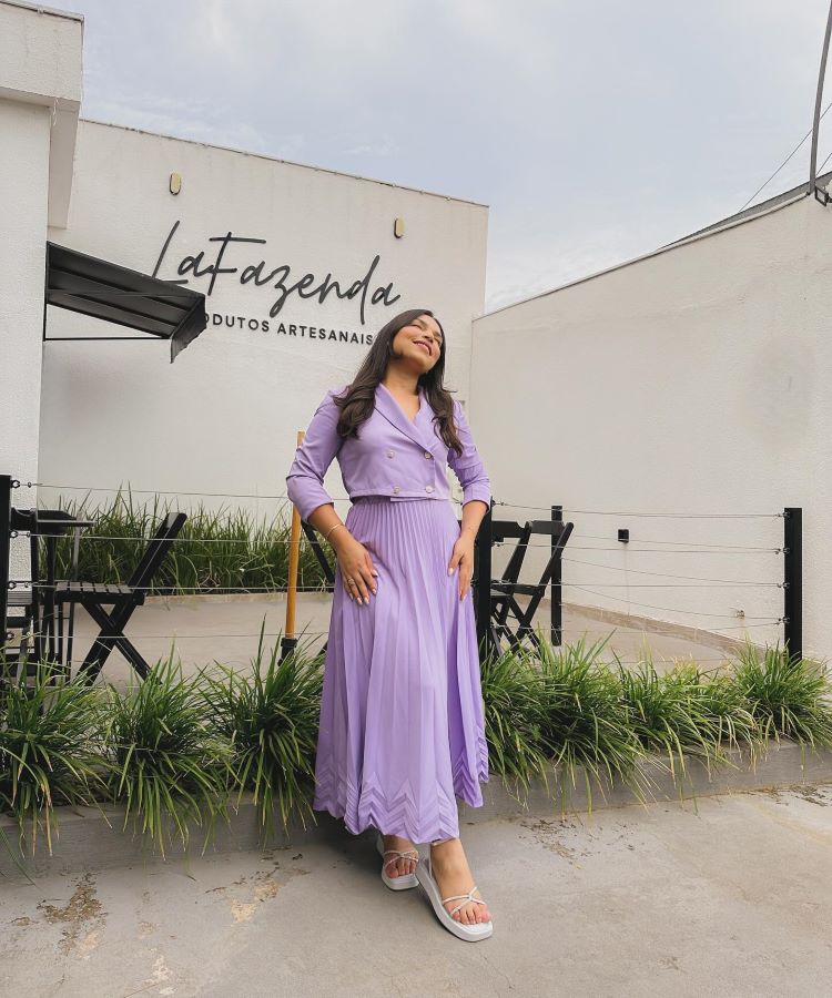 Uma mulher confiante está em pé em frente à loja “Lafazenda Produtos Artesanais”. Ela usa um vestido longo lilás com mangas bufantes, representando a moda evangélica, conhecida por sua elegância e modéstia. Sandálias brancas completam o visual.