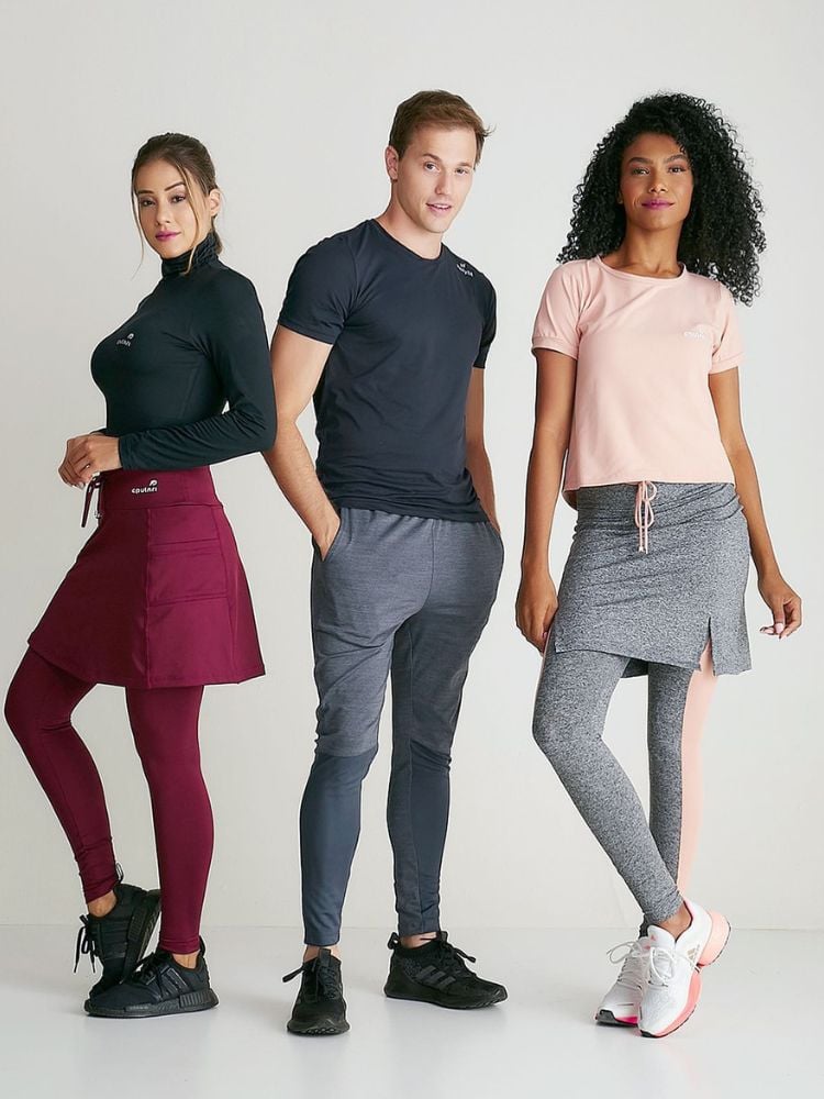 Foto de um homem entre duas mulheres. A da esquerda usa blusa de academia preta manga longa e legging com saia vinho. O homem, ao meio, usa blusa de academia preta e calça cinza. A mulher da direita possui pele negra e usa camiseta solta rosa clara e legging com saia cinzas