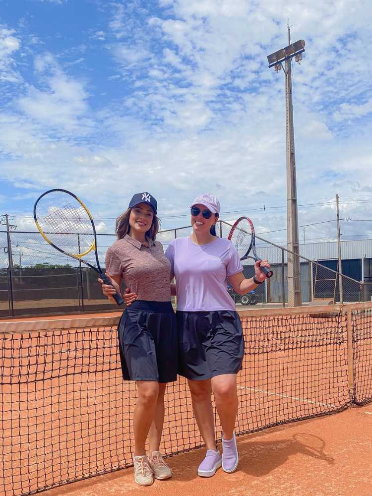 Duas mulheres de pele clara na quadra de tênis com raquete, blusa de academia e shorts saia de tenista até o joelho