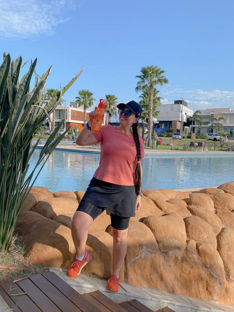 Mulher de pele clara com lago ao fundo, usando blusa solta bege, shorts saia preta quase no joelho e boné preto