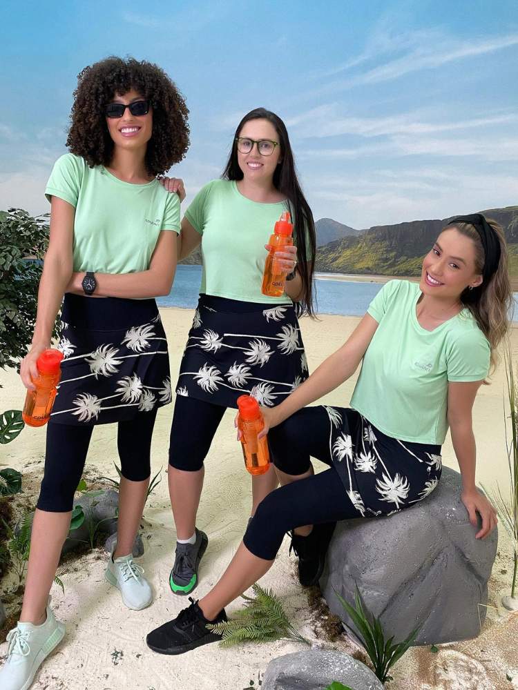 Três mulheres (uma de pele negra e duas de pele clara) usando o mesmo modelo de roupa evangélica para academia: blusa verde e legging com saia pretas, sendo a saia com flores brancas