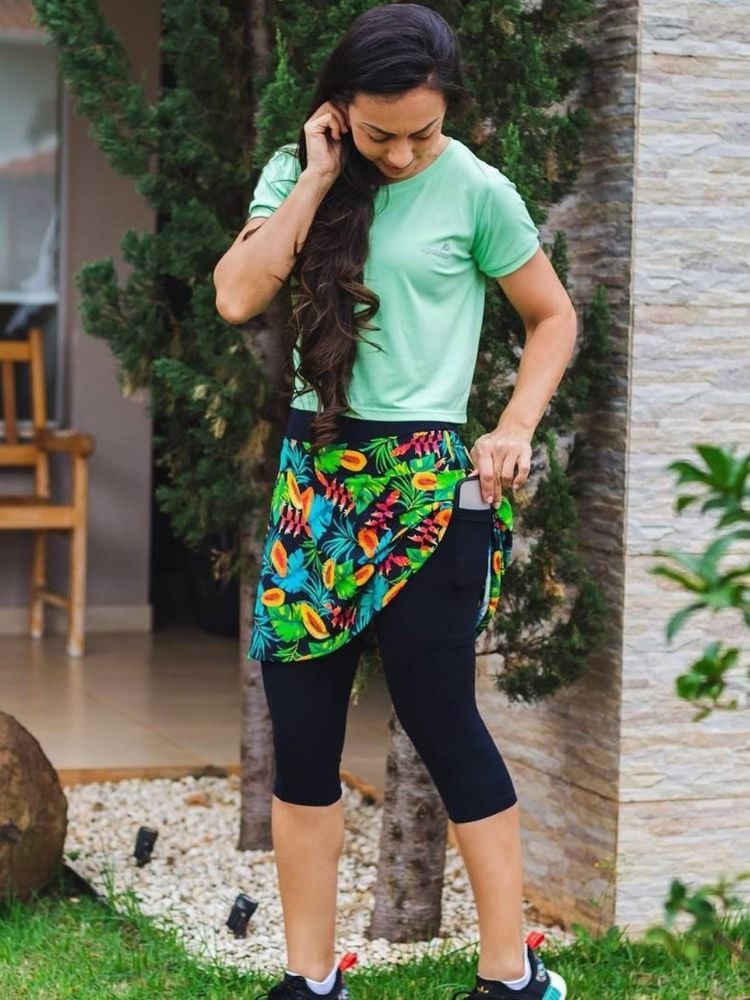 Mulher de pele clara com blusa verde e saia-calça com base preta e saia colorida levantada, mostrando bolso lateral