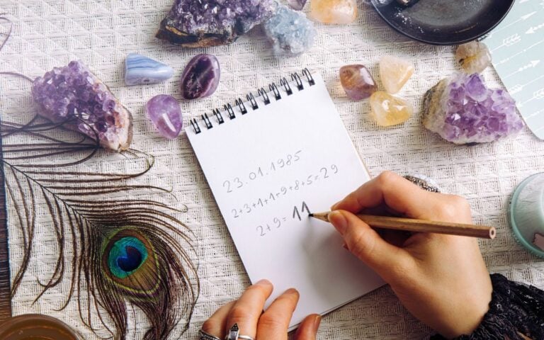 Foto de mãos de mulher calculando número da sorte em um bloco de papel. Ao redor, na mesa, vemos cristais coloridos e uma pena de pavão