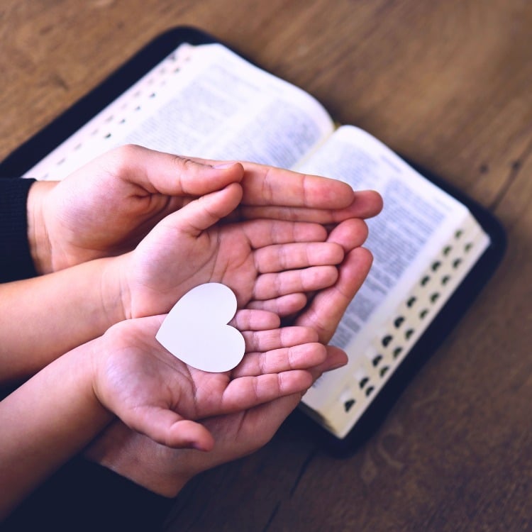 A imagem mostra um par de mãos segurando um coração branco sobre um livro aberto. O livro parece ser uma Bíblia, com páginas finas e texto que aparenta ser de natureza religiosa.