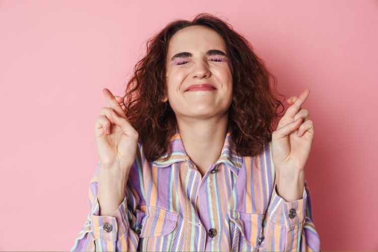 Parede rosa ao fundo, com mulher à frente. Ela tem pele branca, cabelos castanhos, usa camisa listrada e está de olhos fechados e de dedos cruzados, pedindo sorte