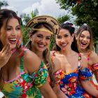 Quatro mulheres sorridentes em roupas festivas e coloridas posam ao ar livre, cercadas por vegetação tropical. Elas usam vestidos com estampas florais vibrantes e acessórios como laços e chapéus. O ambiente transmite alegria e celebração em um cenário tropical iluminado.