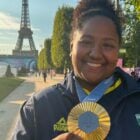 A imagem mostra uma atleta brasileira sorridente, vestindo um uniforme com o brasão do Brasil, segurando uma medalha olímpica dourada. Ao fundo, a Torre Eiffel em Paris, destacando o cenário das Olimpíadas. A medalha exibe o logotipo de Paris 2024, simbolizando a conquista esportiva.