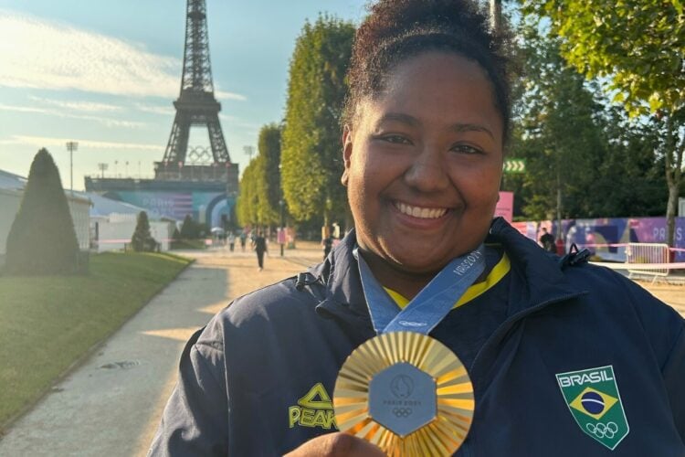 A imagem mostra uma atleta brasileira sorridente, vestindo um uniforme com o brasão do Brasil, segurando uma medalha olímpica dourada. Ao fundo, a Torre Eiffel em Paris, destacando o cenário das Olimpíadas. A medalha exibe o logotipo de Paris 2024, simbolizando a conquista esportiva.