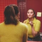 Selena Gomes está em frente a um espelho em um ambiente com paredes revestidas de azulejos vermelhos. Ela está aplicando batom e veste um suéter amarelo felpudo e calças jeans. Refletido no espelho, há um bilhete adesivo com texto escrito à mão.