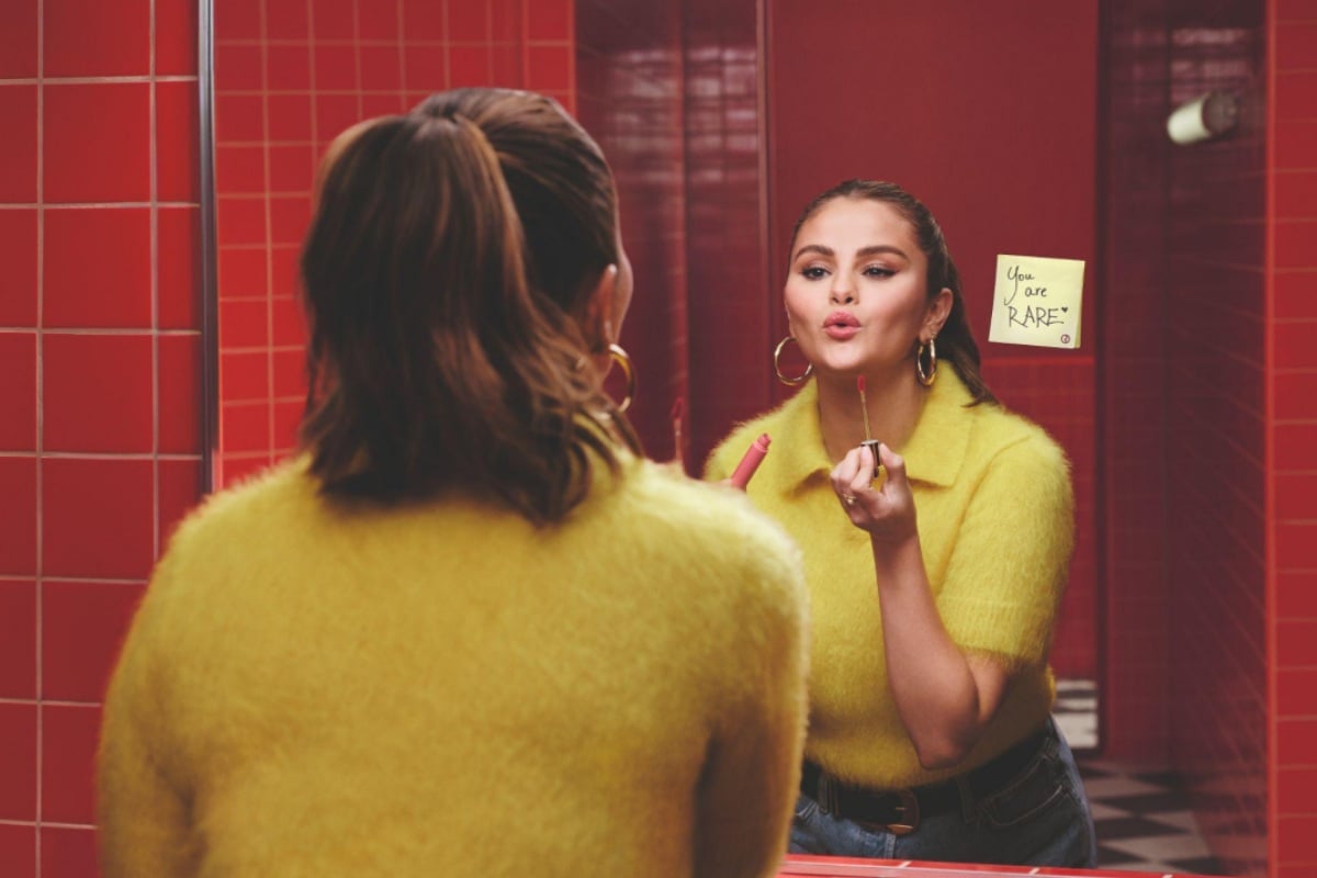 Selena Gomes está em frente a um espelho em um ambiente com paredes revestidas de azulejos vermelhos. Ela está aplicando batom e veste um suéter amarelo felpudo e calças jeans. Refletido no espelho, há um bilhete adesivo com texto escrito à mão.