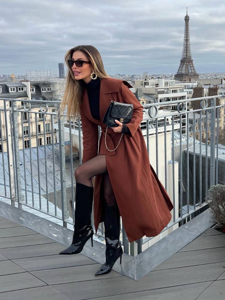 Mulher de pele clara em prédio alto com Torre Eiffel ao fundo, usando meia calça preta, bota cano alto preta, blusa preta e sobretudo marrom