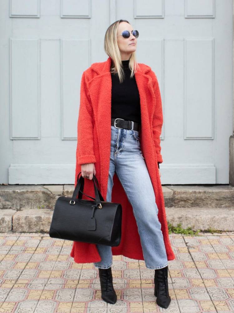 Mulher de pele clara usando sobretudo laranja peludo, blusa preta, bolsa preta, calça jeans e bota preta