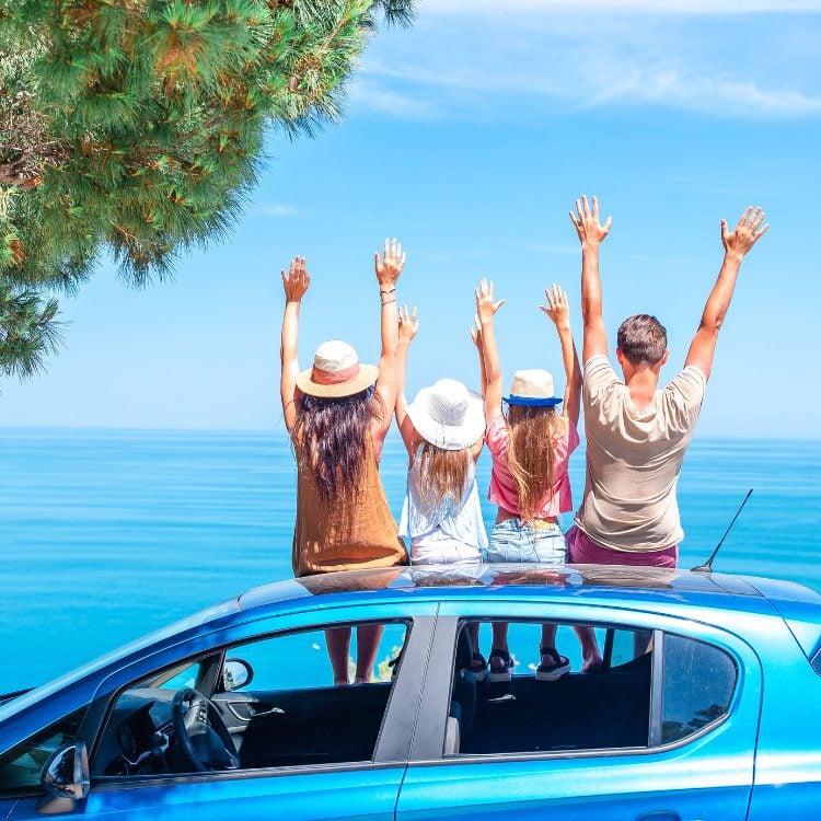 A imagem mostra quatro pessoas de costas, com os braços levantados em celebração, ao lado de um carro azul sob céu claro. Eles estão diante de um vasto oceano azul, sugerindo uma viagem ou passeio familiar. A presença do grupo e a paisagem evocam a ideia de um Dia dos Pais criativo e alegre.