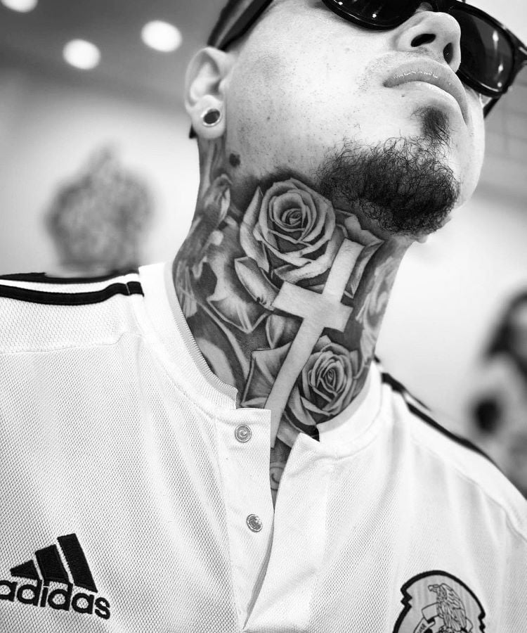 A imagem mostra uma pessoa de frente com a cabeça inclinada para trás. Ela tem uma tatuagem no pescoço de grandes rosas e uma cruz central. A pessoa usa óculos escuros, brincos e uma camisa branca da Adidas.