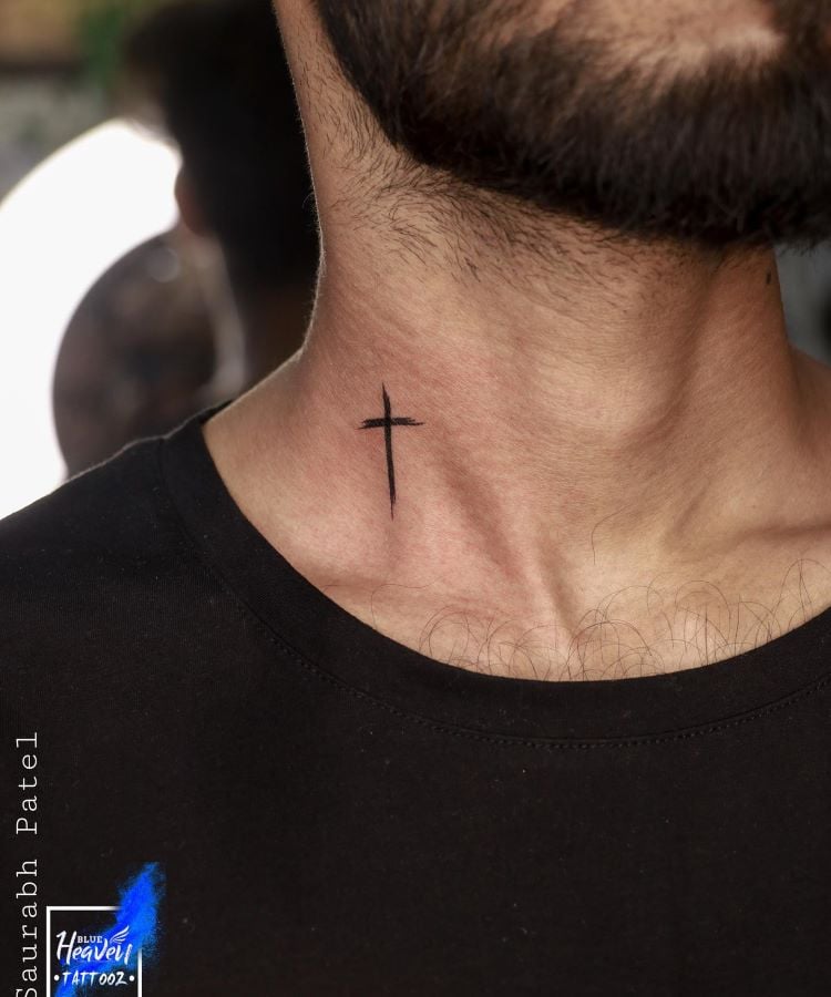 A imagem mostra uma pessoa com barba curta e cabelo escuro, usando uma camiseta preta. Ela tem uma tatuagem no pescoço de uma cruz simples e preta, localizada na parte frontal do pescoço, logo abaixo da linha do queixo. A tatuagem é minimalista e centralizada.