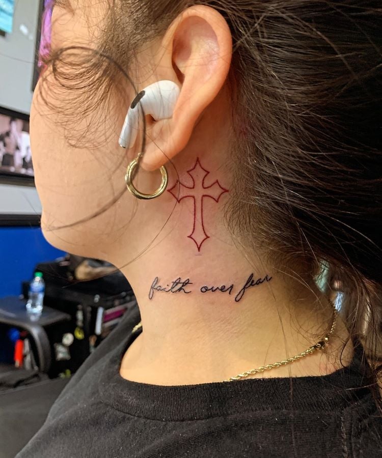 A imagem mostra uma pessoa de perfil com cabelo preso. Ela tem uma tatuagem no pescoço de uma cruz vermelha estilizada localizada logo abaixo da orelha. Abaixo da cruz, há uma frase em preto que diz "faith over fear" (fé sobre medo). A pessoa usa um brinco dourado grande e um fone de ouvido.