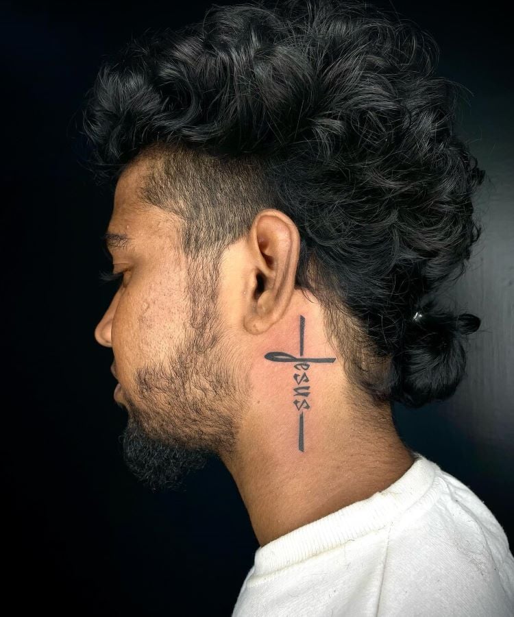 A imagem mostra uma pessoa de perfil com cabelo encaracolado preso em um coque. Ela tem uma tatuagem no pescoço que consiste na palavra "Jesus" escrita verticalmente, formando uma cruz. A pessoa usa uma camiseta branca.