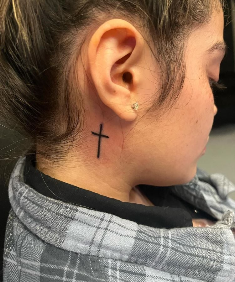 A imagem mostra uma pessoa de perfil com cabelo preso. Ela tem uma tatuagem no pescoço de uma pequena cruz preta localizada atrás da orelha. A pessoa usa um brinco pequeno e está vestida com uma camisa xadrez cinza e preta.