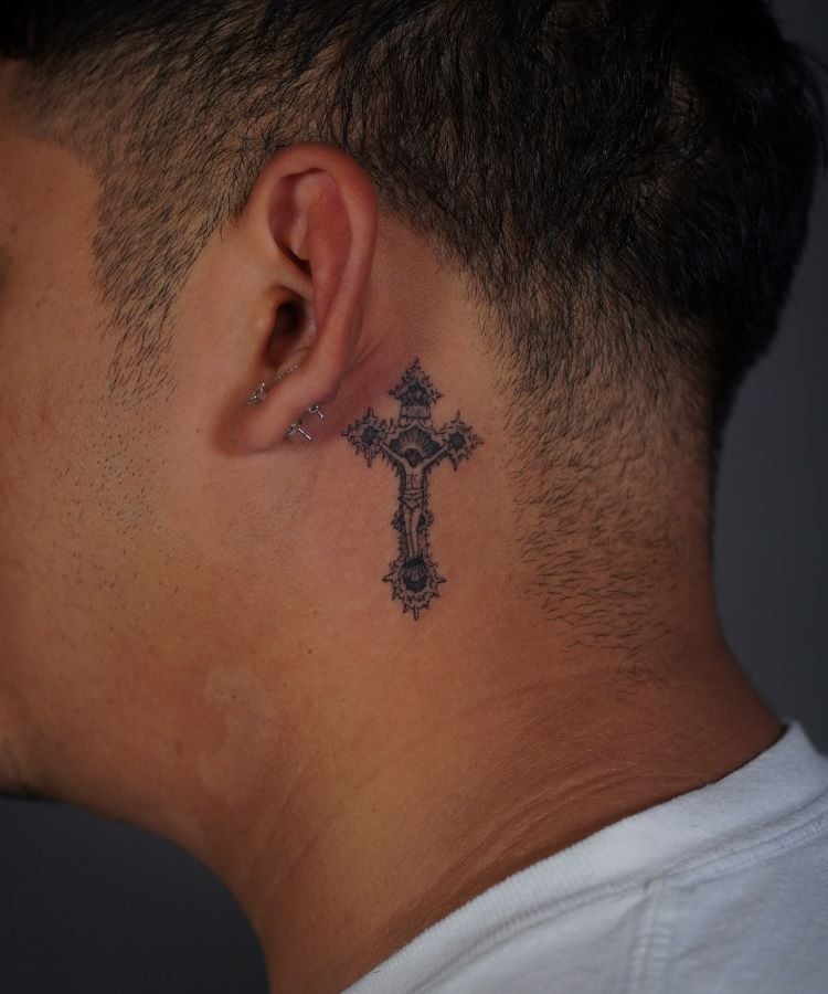 A imagem mostra uma pessoa de perfil com cabelo curto e escuro. Ela tem uma tatuagem no pescoço de uma cruz detalhada em preto, localizada logo abaixo da orelha. A pessoa usa uma camiseta branca e pequenos piercings na orelha.