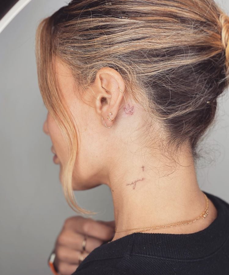 A imagem mostra uma pessoa de perfil com cabelo loiro preso em um coque. Ela tem uma tatuagem no pescoço de uma pequena pombinha atrás da orelha e a palavra "imagine" escrita em vermelho com uma pequena cruz preta. A pessoa usa brincos pequenos e uma corrente dourada.