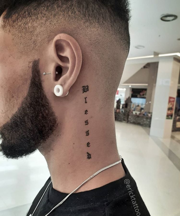 A imagem mostra uma pessoa de perfil com cabelo curto e barba. Ela tem uma tatuagem no pescoço com a palavra "Blessed" escrita verticalmente em letras pretas. A tatuagem está localizada na lateral do pescoço, perto da orelha. A pessoa também usa um alargador branco na orelha e um brinco de argola.