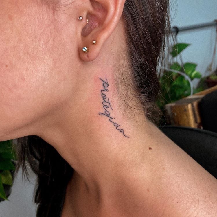 A imagem mostra uma pessoa de perfil com cabelo preso. Ela tem uma tatuagem no pescoço com a palavra "protegida" escrita em letras cursivas pretas. A tatuagem está localizada na lateral do pescoço, perto da orelha, onde a pessoa também usa vários brincos pequenos.