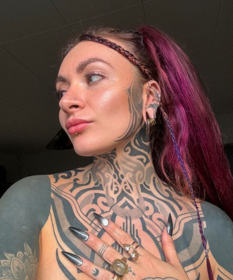 A imagem mostra uma pessoa de perfil com cabelo roxo preso em um rabo de cavalo. Ela tem uma tatuagem no pescoço e no peito de padrões tribais em preto, que se estendem até o rosto. A tatuagem é complexa e cobre grande parte da pele visível.