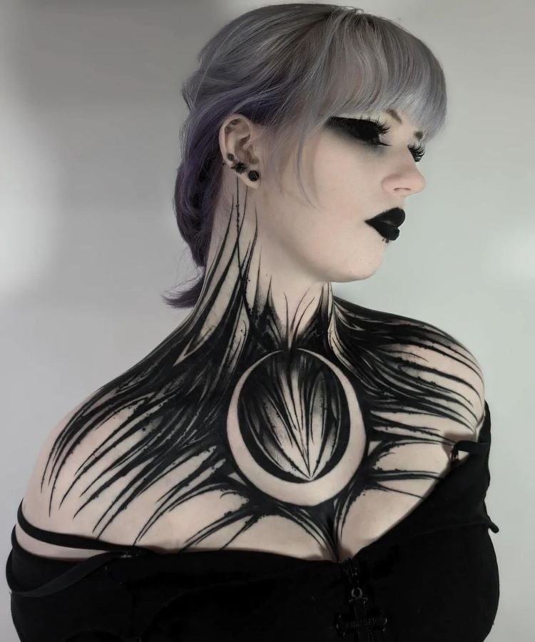 A imagem mostra uma pessoa com cabelo cinza claro e maquiagem escura. Ela tem uma tatuagem no pescoço que se estende até o peito, com um design abstrato em preto, lembrando penas ou garras, e um círculo central formando uma lua. A tatuagem cobre o pescoço e a parte superior do peito.