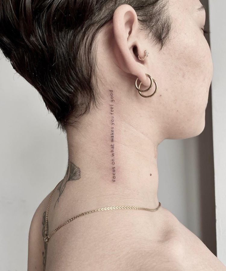 A imagem mostra uma pessoa de perfil com uma tatuagem no pescoço que consiste em uma frase escrita verticalmente. A frase diz "focus on what makes you feel good". A pessoa tem cabelo curto, usa dois brincos de argola dourados e uma corrente dourada no pescoço.