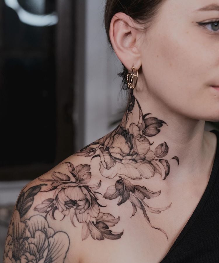 A imagem mostra uma pessoa de perfil com cabelo castanho preso. Ela tem uma tatuagem no pescoço de flores detalhadas em preto e cinza, que se estendem até o ombro. A tatuagem é delicada e realista, cobrindo a lateral do pescoço e parte do ombro.